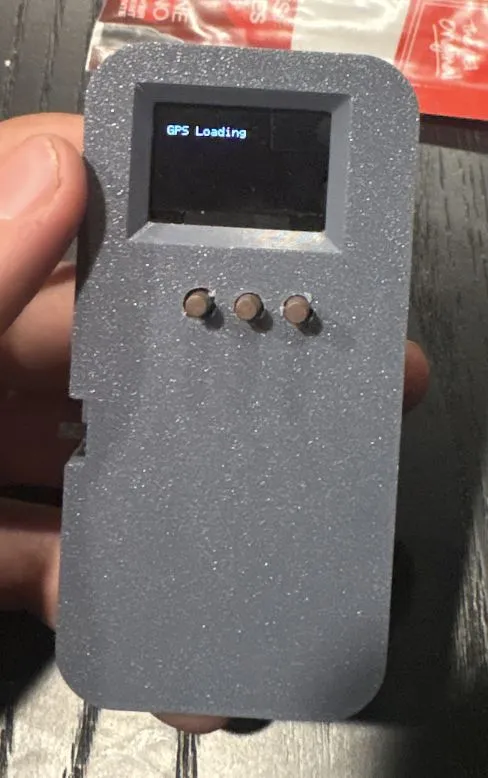 Handheld range finder in a gray 3D-printed enclosure, held in one hand. The OLED screen reads 'GPS Loading' above three round buttons.