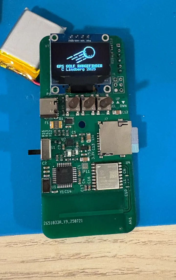 Assembled Rev 2 range finder board on a blue mat: the OLED screen shows a golf ball graphic and 'GPS GOLF RANGEFINDER, C Lindberg 2025' above three push buttons, a USB-C port, a microSD card slot and the GNSS module, with a small LiPo battery beside it.