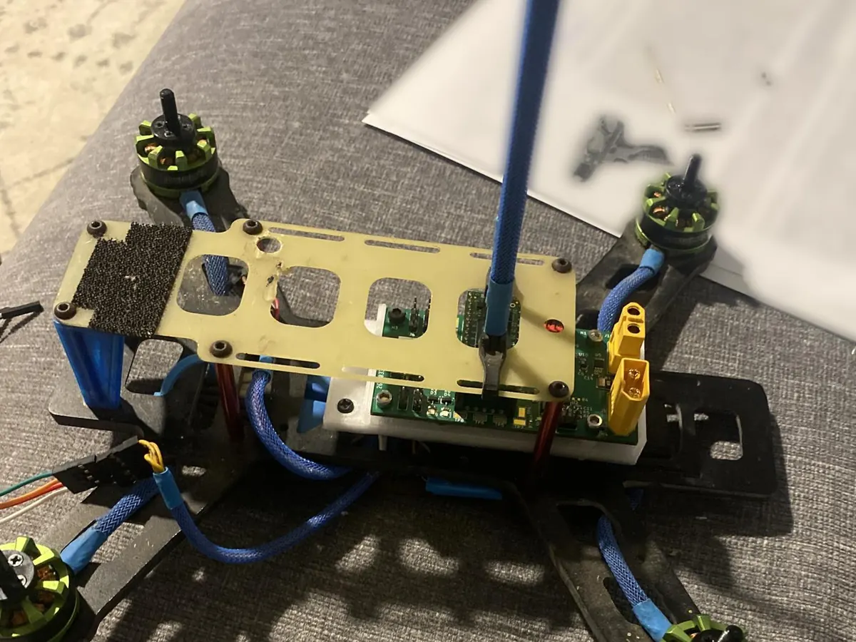 Quadcopter frame with the flight controller board mounted under a top plate in a white holder, yellow XT60 connectors on one side, a vertical receiver antenna, and green brushless motors on the carbon-fiber arms.