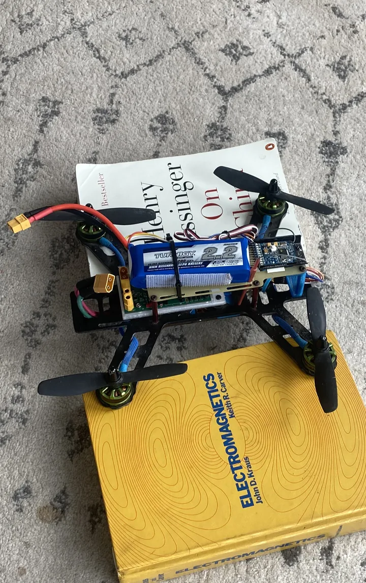 Small carbon-fiber quadcopter with four black propellers and green motors, a blue 2.2 Ah LiPo battery strapped on top and yellow XT60 connectors, resting on two books on a patterned carpet.